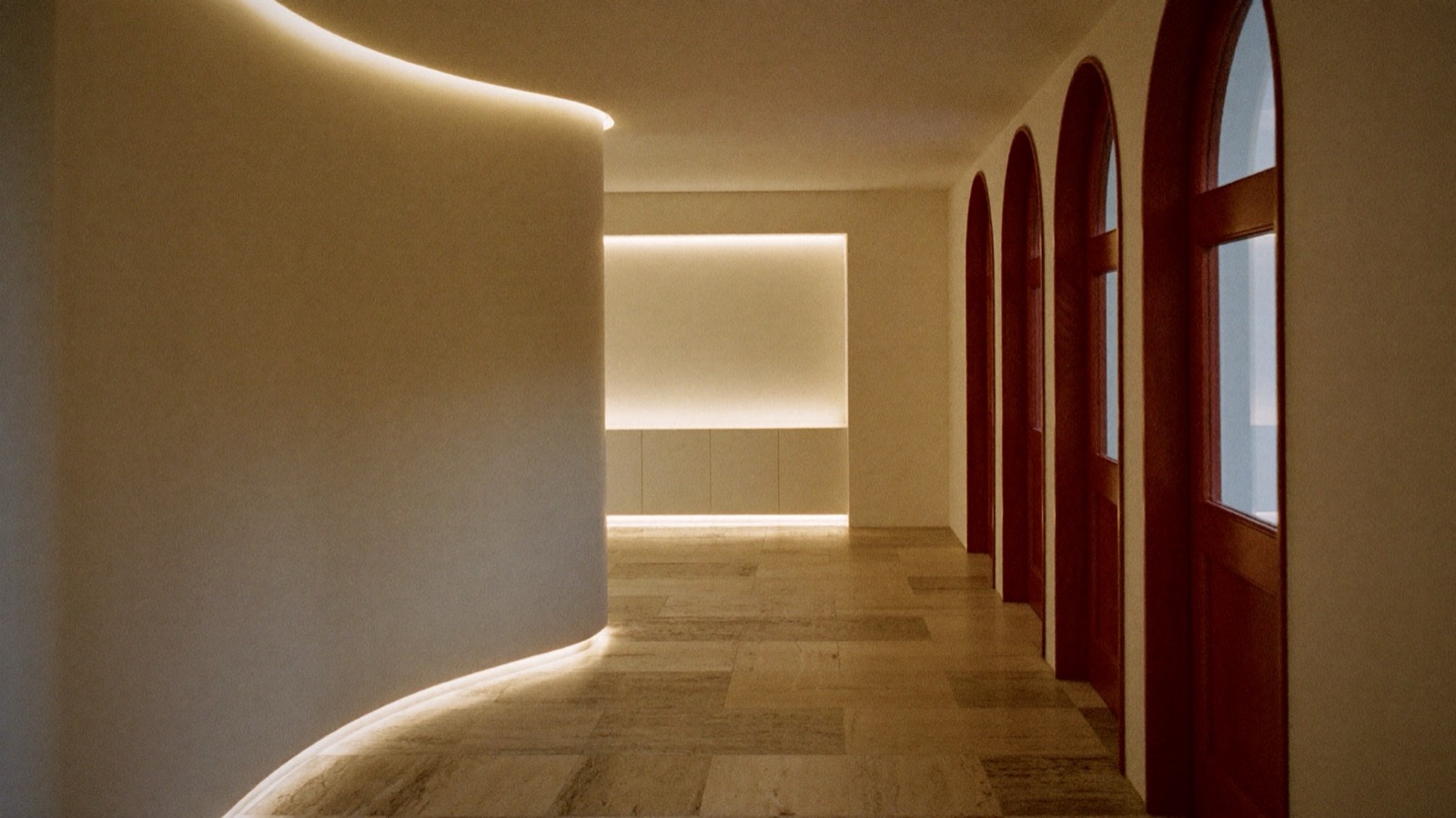 A curved cream plaster corridor with a warm lit edge, arched oxblood doors and a travertine floor.