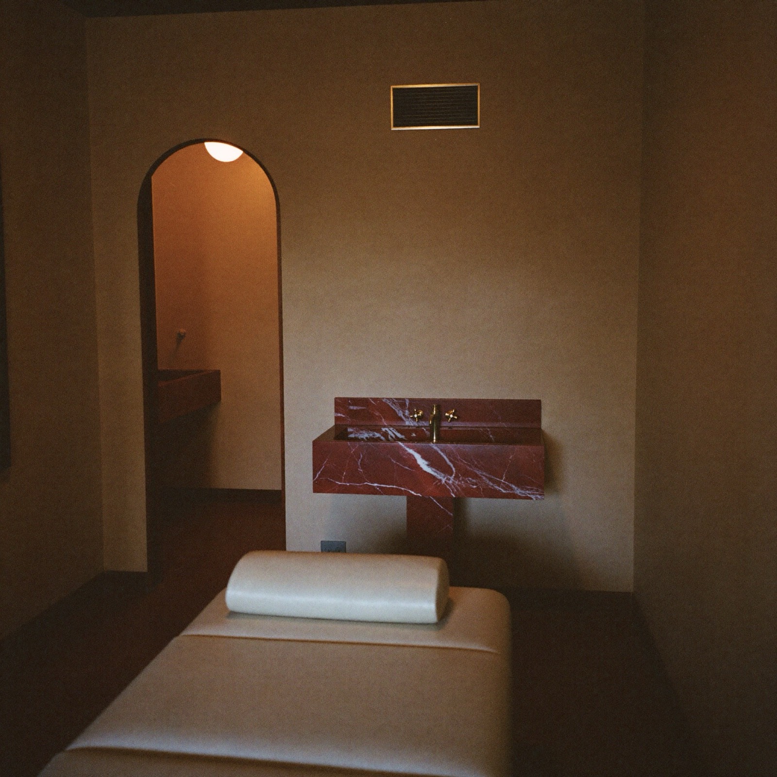 A treatment room with a cream leather bed, a red marble basin and a lit arched doorway.